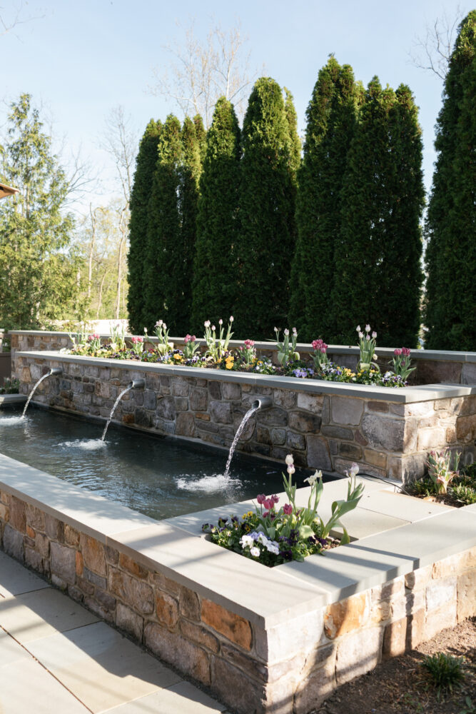Stone rectangular water feature built into a stone planter with three spouts bordered by blooming tulips and other flowers and tall evergreen trees in the background