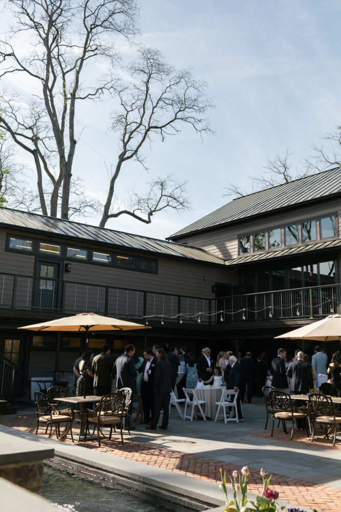 Guests in formal attire mingle on a brick patio courtyard outside a modern brown venue with string lights and umbrellas