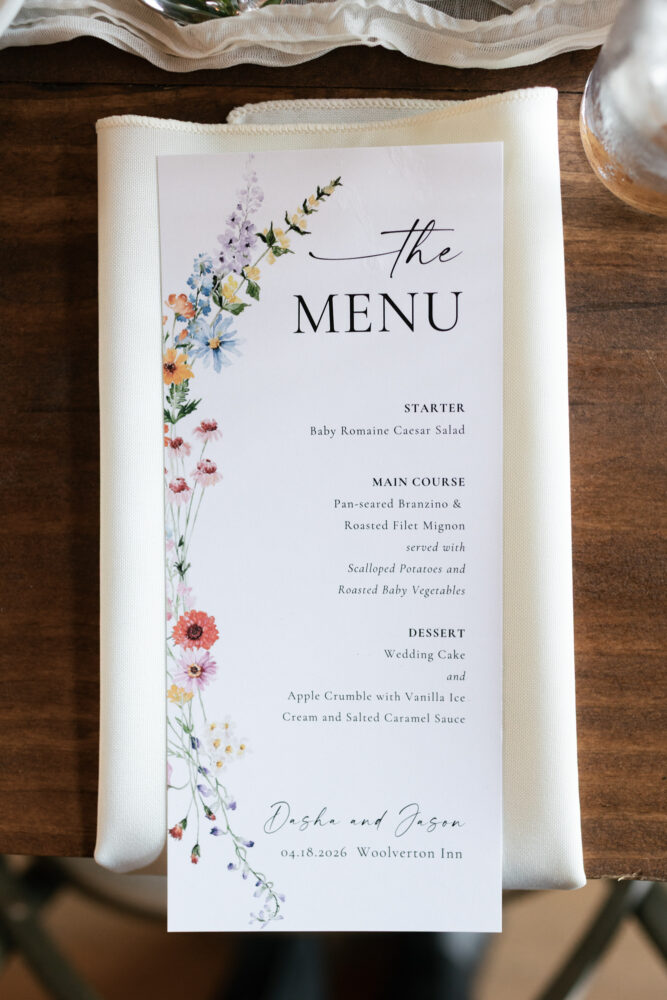 Wedding menu card with colorful flower border on the left placed on a folded white napkin on a wooden table