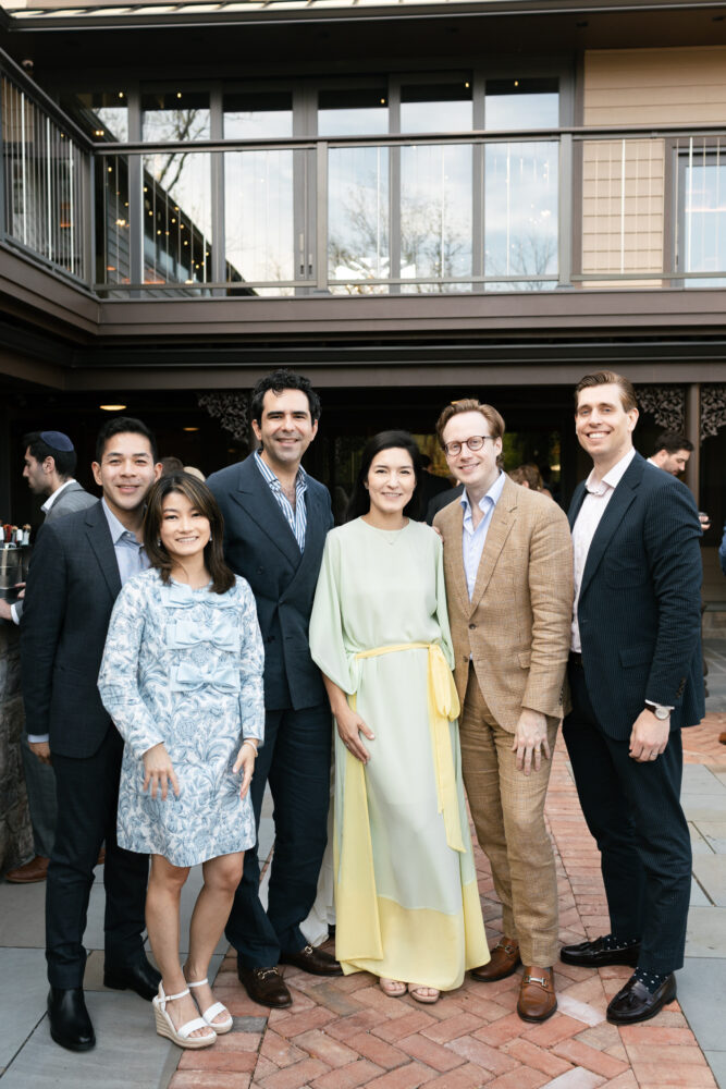 Six well dressed adults posing together at an outdoor social event with a brick plaza and building in the background smiling for the photo