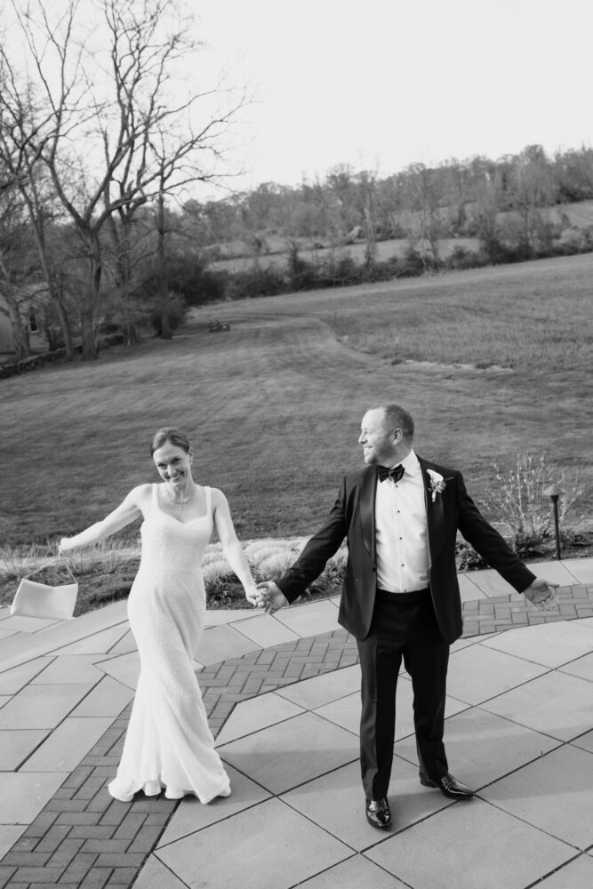 Bride in a white wedding dress and groom in a black tuxedo hold hands while walking on a stone terrace outdoors in a black and white scene