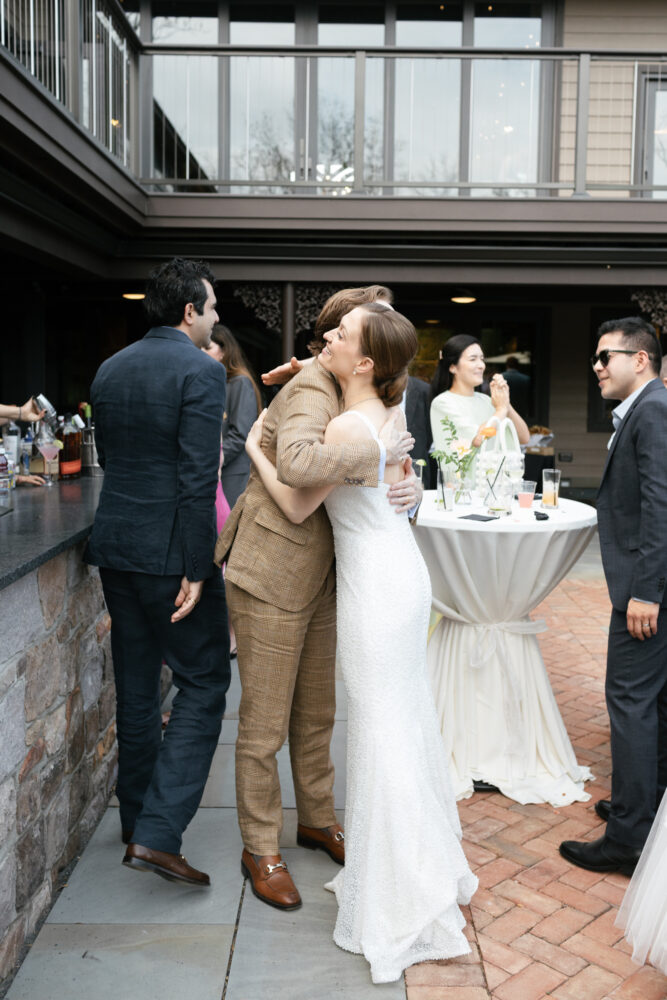 Bride in a white gown hugs a man in a tan plaid suit at an outdoor wedding reception by a bar