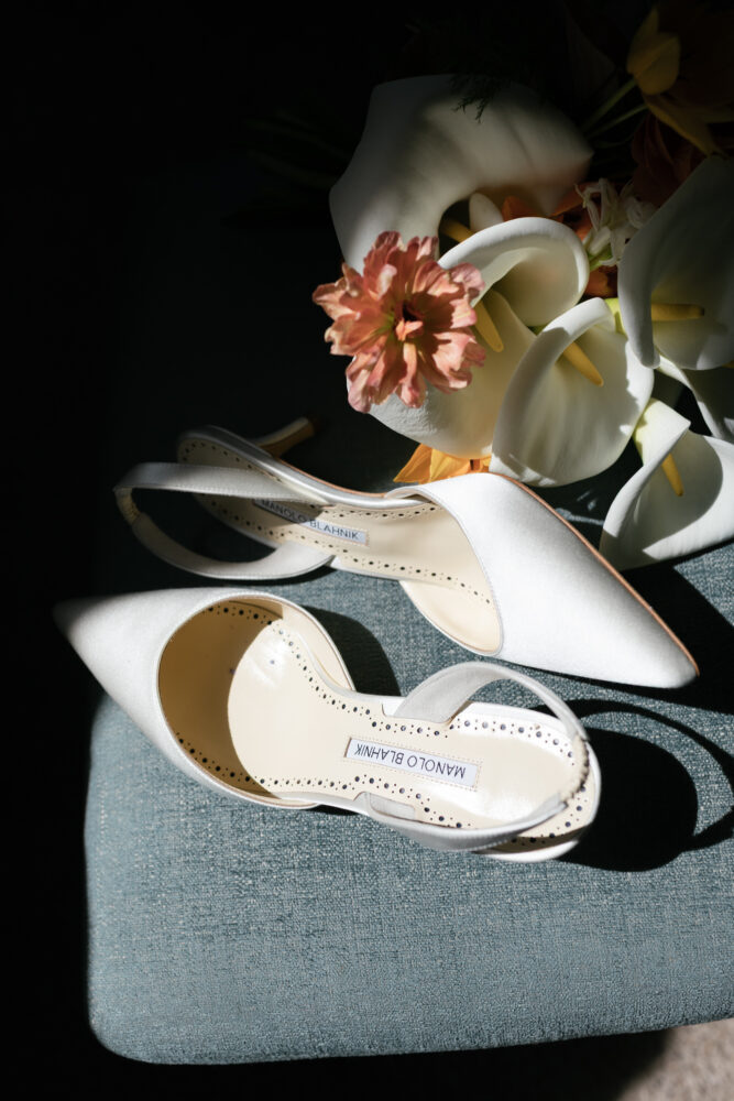 White satin high heels with a bouquet of calla lilies and a pink flower on a teal fabric surface; Manolo Blahnik label visible inside one shoe.