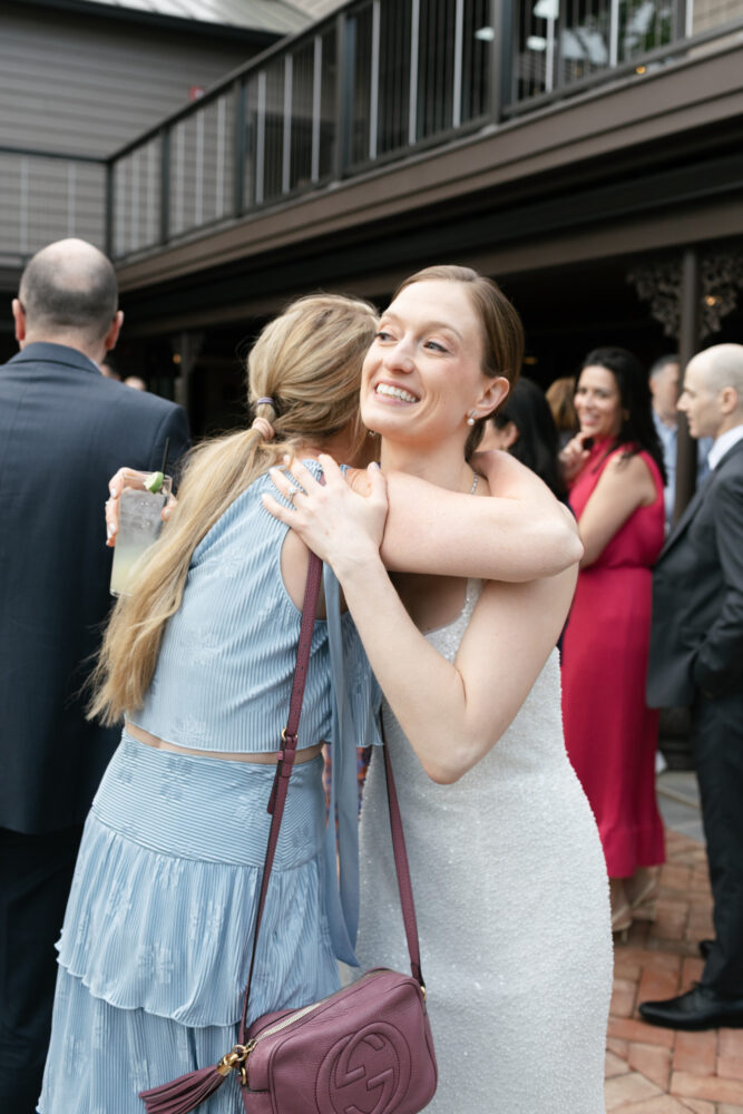 Bride in a beaded white gown hugs a friend in a light blue dress at an outdoor wedding reception the friend holding a cocktail and a purple purse