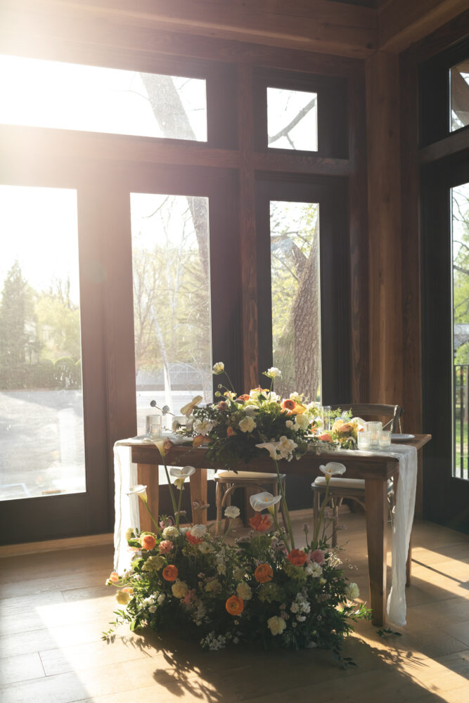 Sunlit dining table decorated with a large flower arrangement and smaller centerpieces inside a wooden framed room