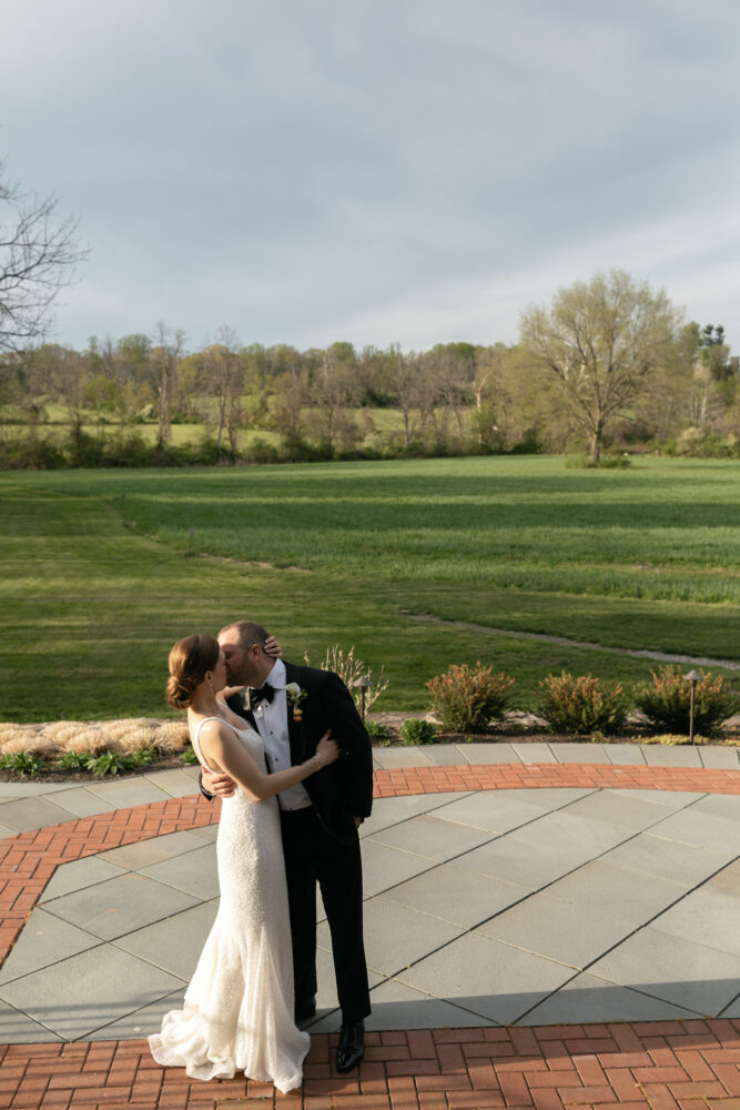 Bride in a white wedding gown and groom in a black tuxedo kiss on a stone and brick patio with a green lawn behind them trees in the distance