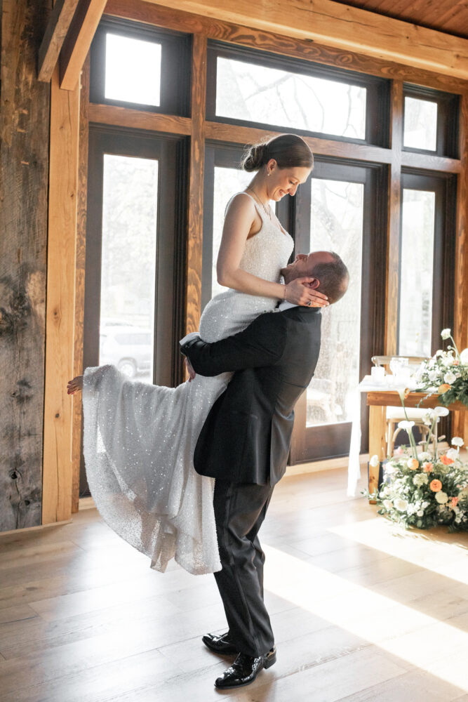 Bride in a sparkly white wedding gown being lifted by groom in a black suit indoors smiling at each other amid lantern light
