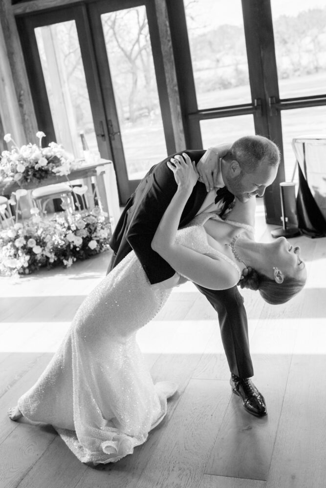 Bride and groom in a dip pose during a wedding dance indoors with floral decor in the background black and white