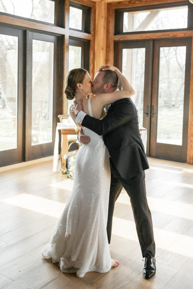Bride in a sparkling white wedding gown kisses and hugs her groom in a sunlit wooden room with large windows behind them