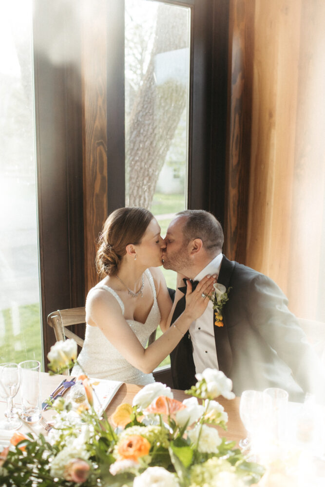 Bride and groom kissing at their wedding reception seated beside a floral centerpiece by a large window