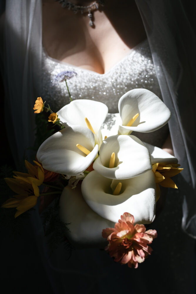 Brides bouquet featuring white calla lilies and small yellow blooms held against a glittering wedding dress