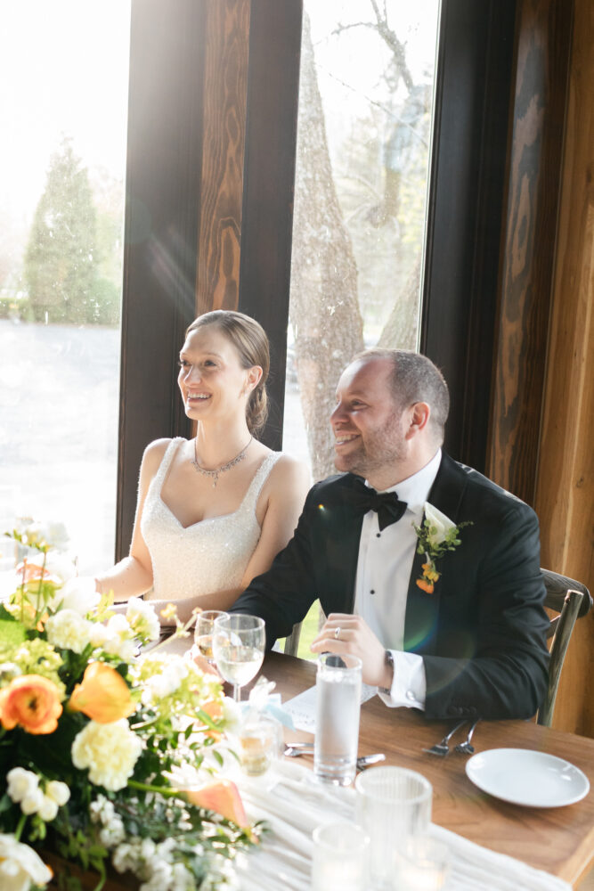 Bride and groom seated at a sunlit wedding reception table smiling with a floral centerpiece and glassware nearby