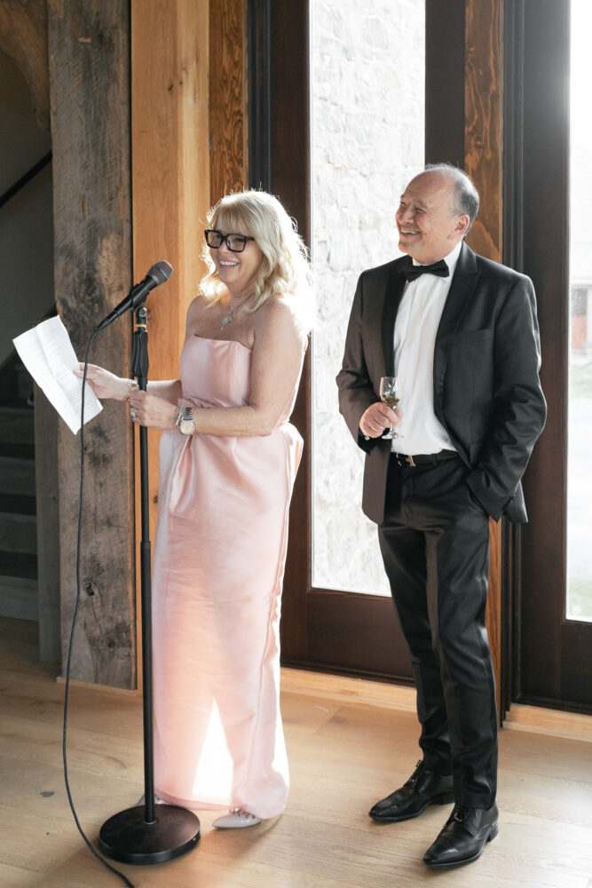 Woman in a pink strapless gown speaks into a microphone holding papers as a smiling man in a tuxedo stands nearby
