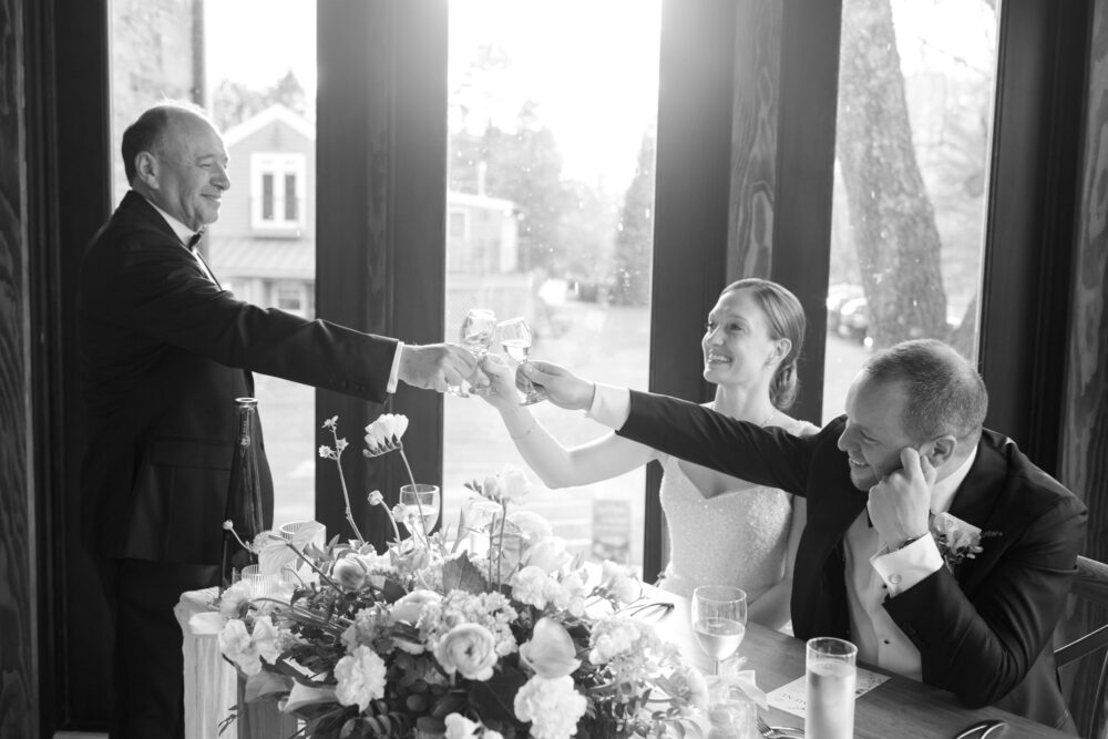 Wedding toast bride and groom raise glasses with an older man as they sit at a flower filled table in a bright room