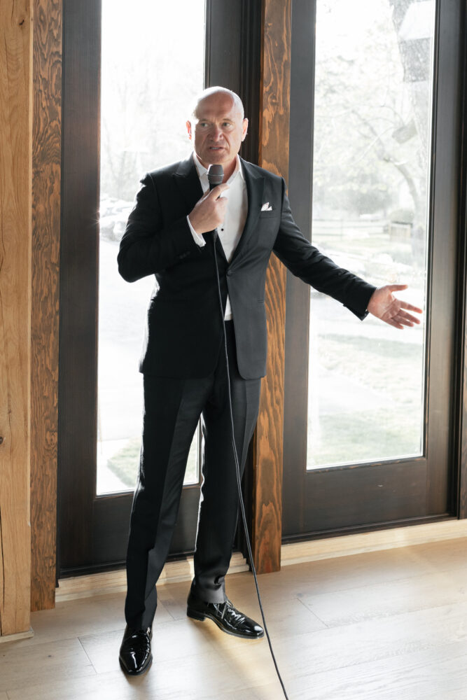 Man in a black suit and white shirt speaks into a handheld microphone gesturing with his other hand indoors by large windows with wooden trim
