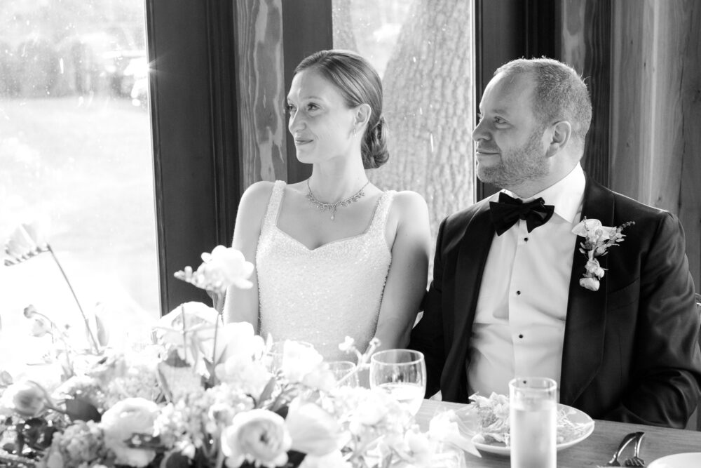 Bride and groom in formal wear seated at a flower adorned table during a wedding reception looking to the right with smiles