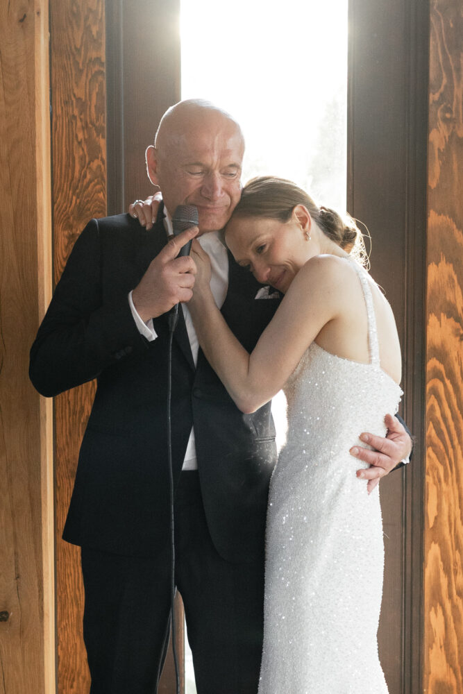 Bride in a sparkling white gown leans on an older man in a suit as he holds a microphone singing together by a wooden doorway
