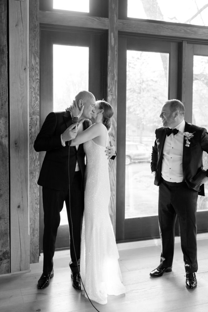 Bride in a sequined gown kisses her father as a man in a tuxedo grins nearby in a rustic window filled venue