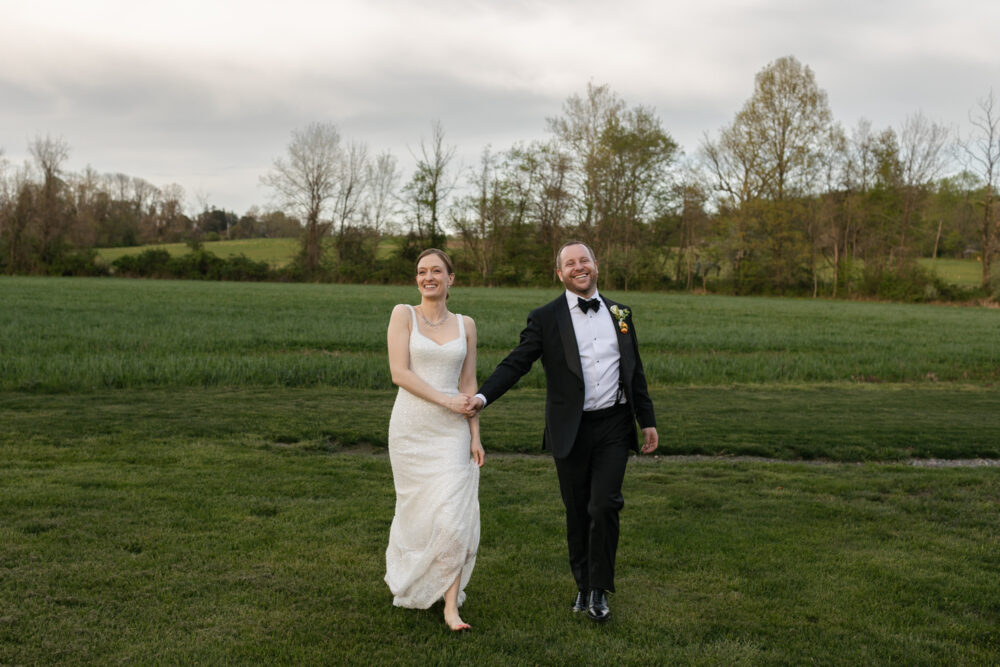 A bride in a white wedding gown and a groom in a black tuxedo walk hand in hand across a grassy field smiling in a rural outdoor setting