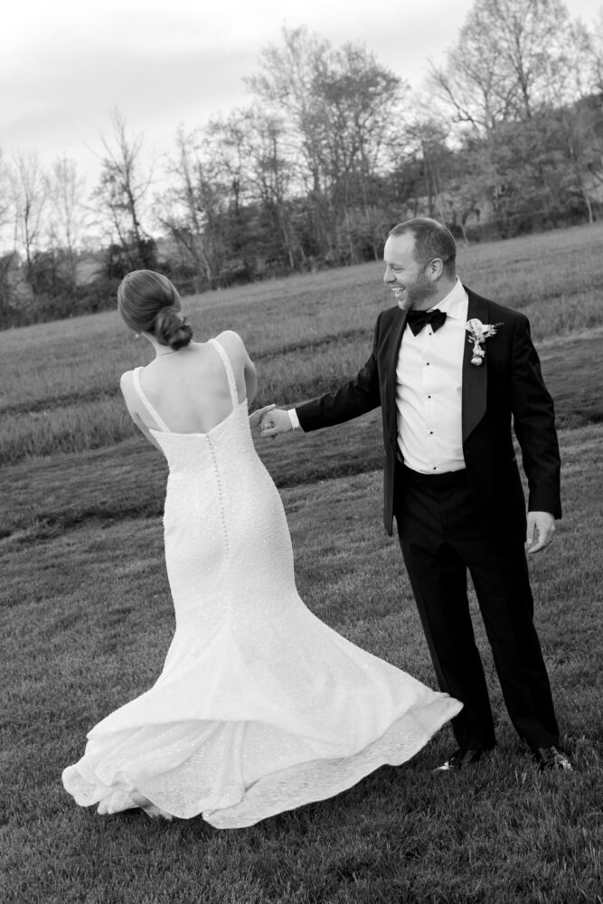 Bride and groom in formal attire hold hands and dance on a grassy field with trees in the background blackandwhite photo