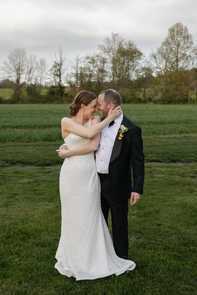 Bride in a sparkling white gown embraces a groom in a black tuxedo in a green field with trees in the background smiling at each other