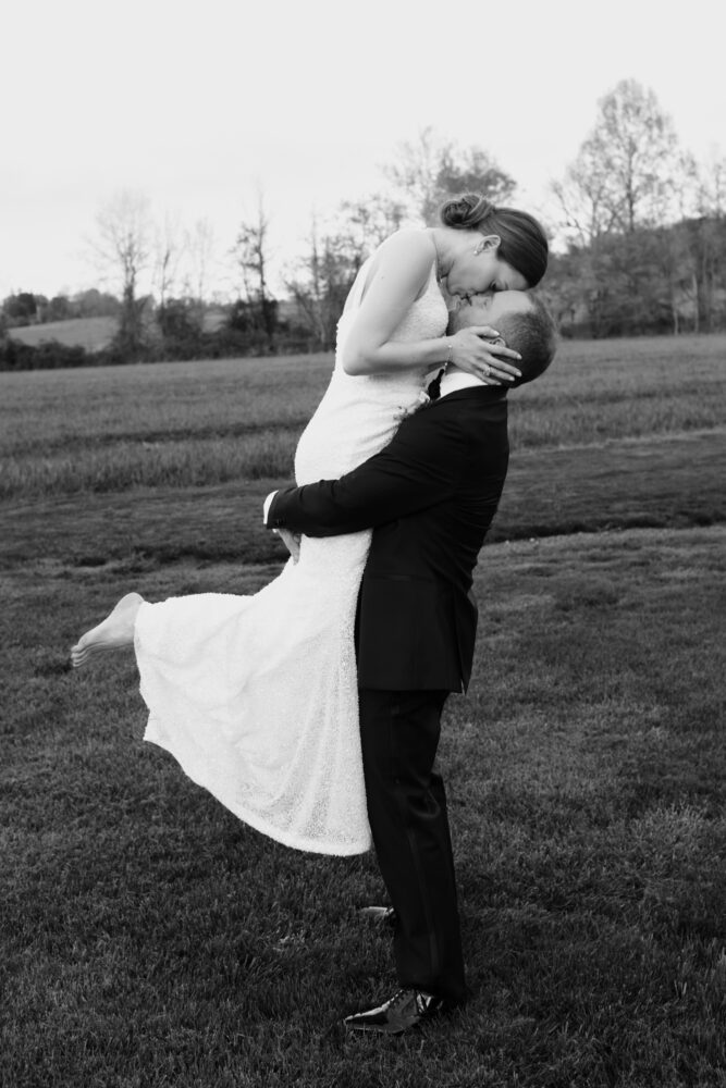 Bride in a white wedding dress is lifted and kissed by her groom in a grassy field outdoors