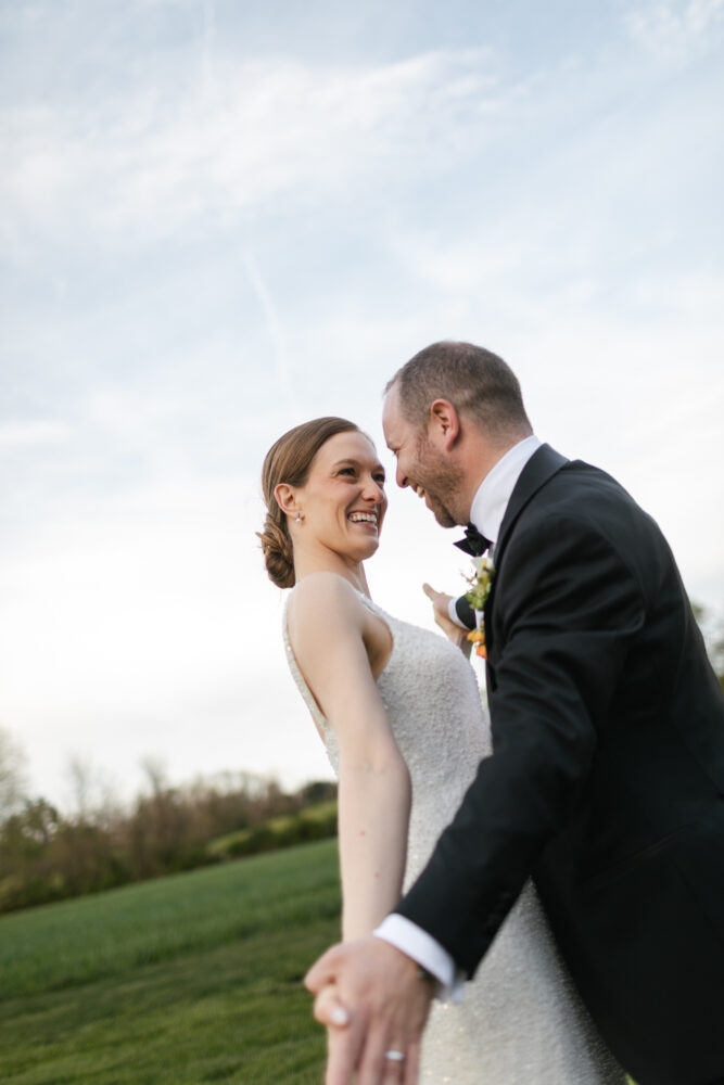 Bride and groom laugh while holding hands in a grassy outdoor setting she wears a beaded white gown he in a black suit and bow tie