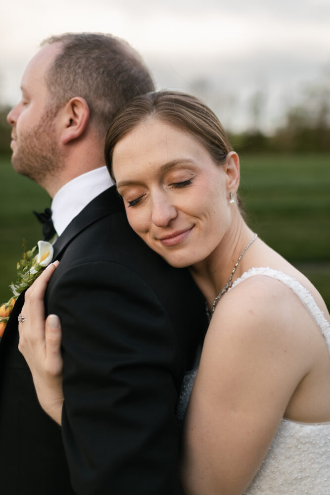 Bride rests her head on her grooms shoulder with a gentle smile outdoors