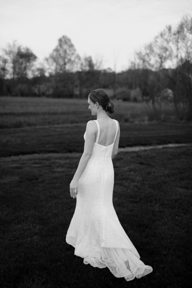 Bride in a sparkling white wedding gown walking away across a grassy field with trees in the distance back view