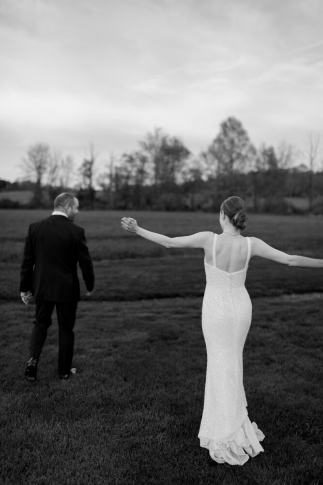 Back view of a bride in a sparkling white dress with arms outstretched walking across a grassy field beside a man in a suit