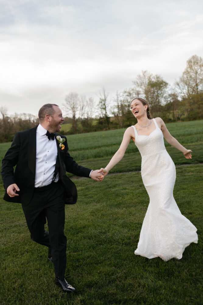 Bride in a beaded white wedding dress and a black tie groom run hand in hand across a green field laughing