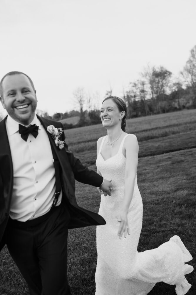 Black and white photo of a smiling bride in a beaded wedding gown and a groom in a tuxedo laughing as they walk hand in hand across a grassy field