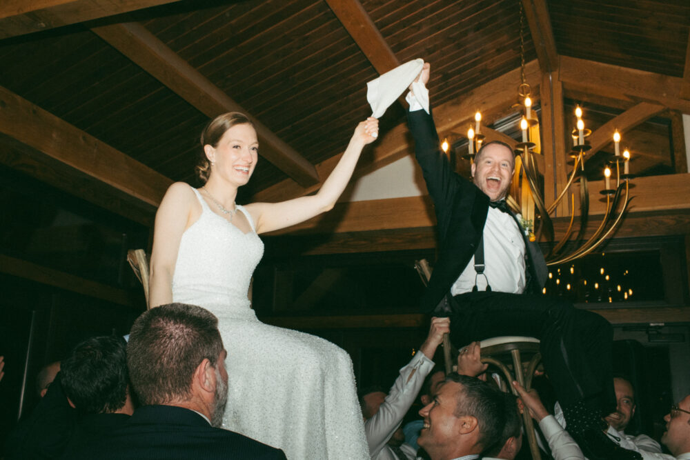Bride and groom are lifted on chairs by guests celebrating and waving napkins during a wedding reception Hora style