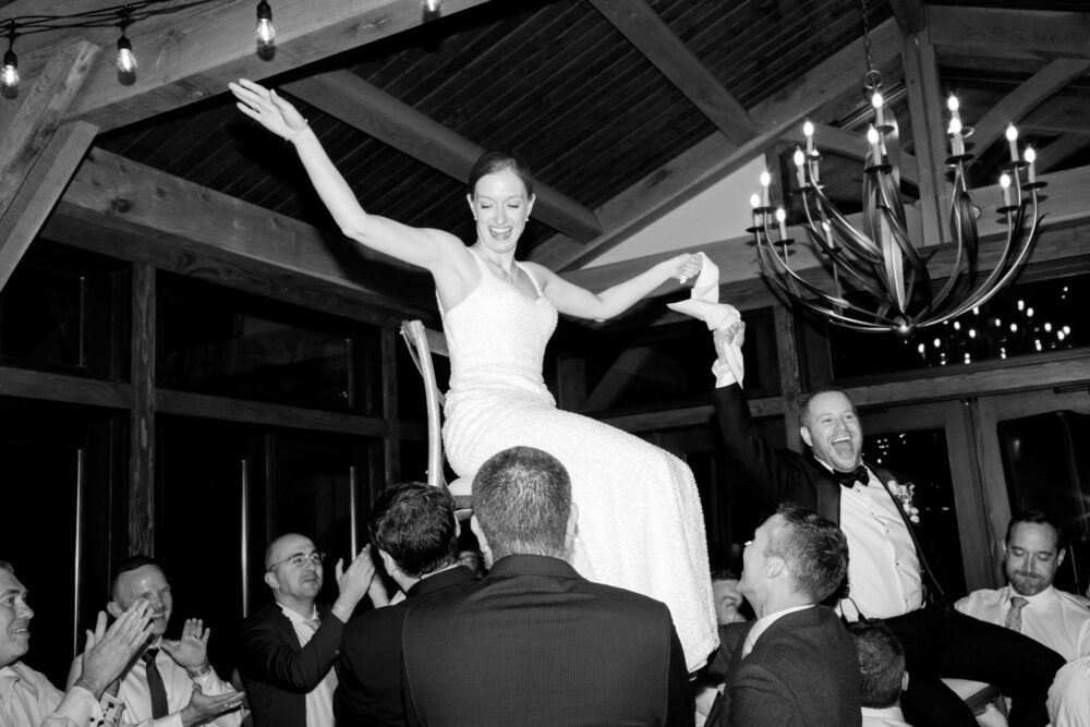 Bride in a white wedding gown sits on shoulders above a crowd arms raised in celebration as guests cheer at a wedding reception in a wood beamed venue with a chandelier above