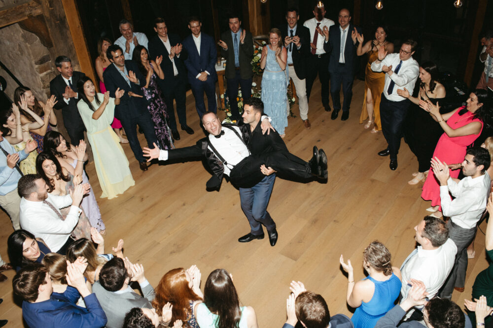 Guests in formal attire form a circle on a wooden dance floor as one man carries another in a playful lift during a wedding celebration