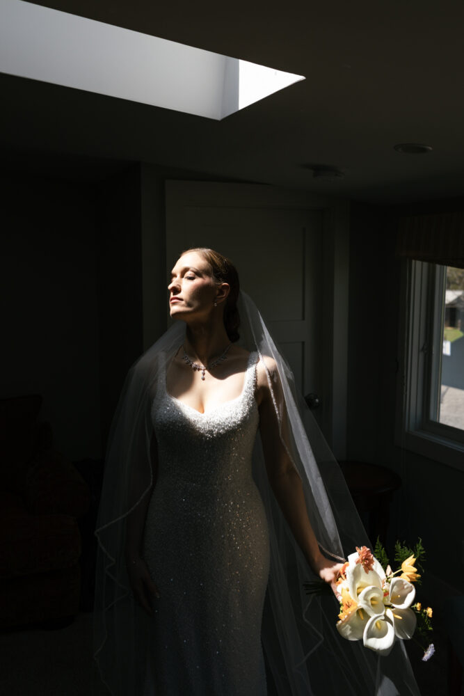 Bride in a sparkling beaded wedding gown and veil stands in a dim room, head turned toward a skylight, holding a white-and-orange bouquet.