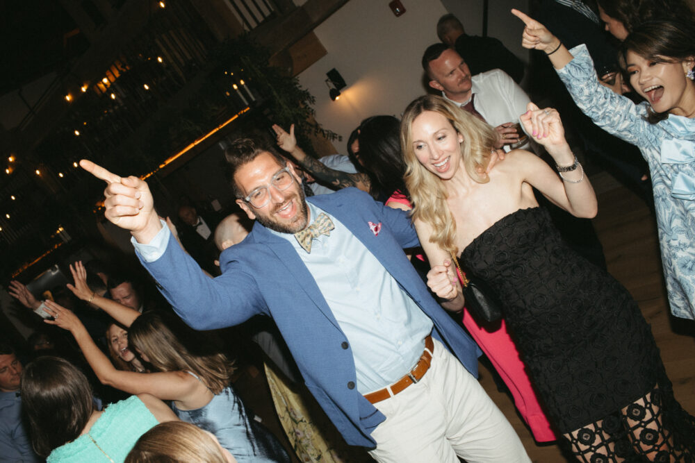 Group of friends dancing at a party man in a blue blazer and bow tie pointing with both hands while smiling at the camera