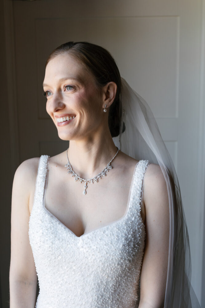 Bride in a beaded wedding gown smiling softly wearing a veil and diamond necklace standing by a doorway