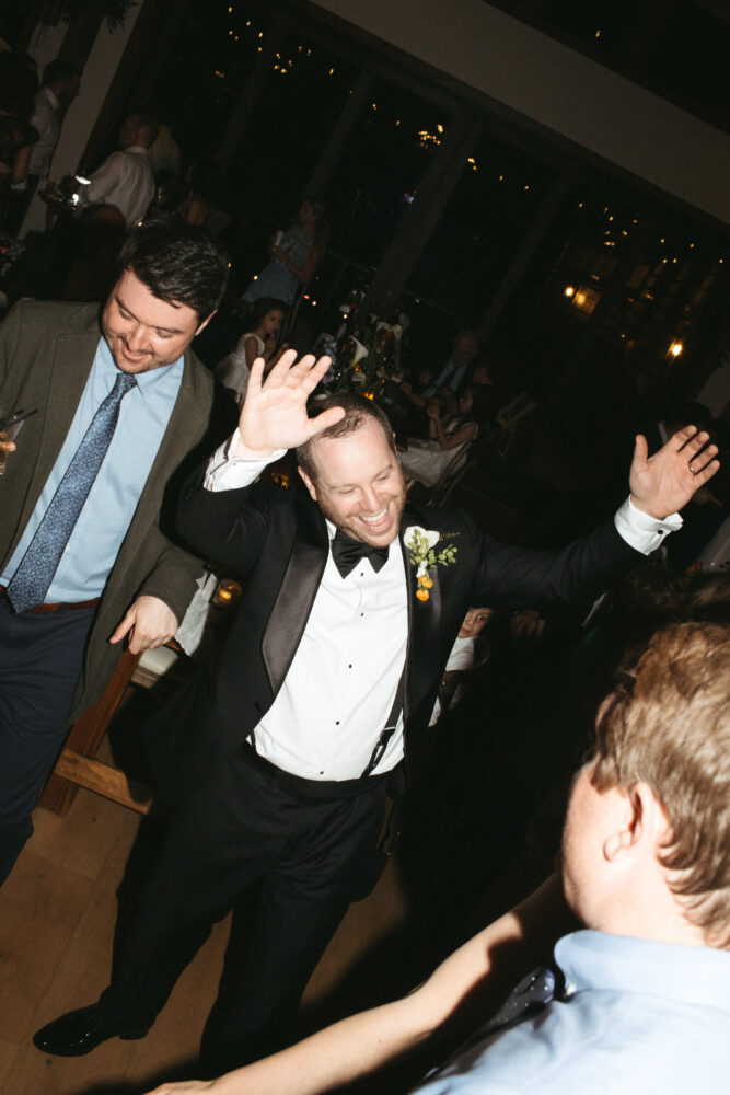 Groom in a black tuxedo and bow tie raises his hands and smiles while dancing at a wedding reception