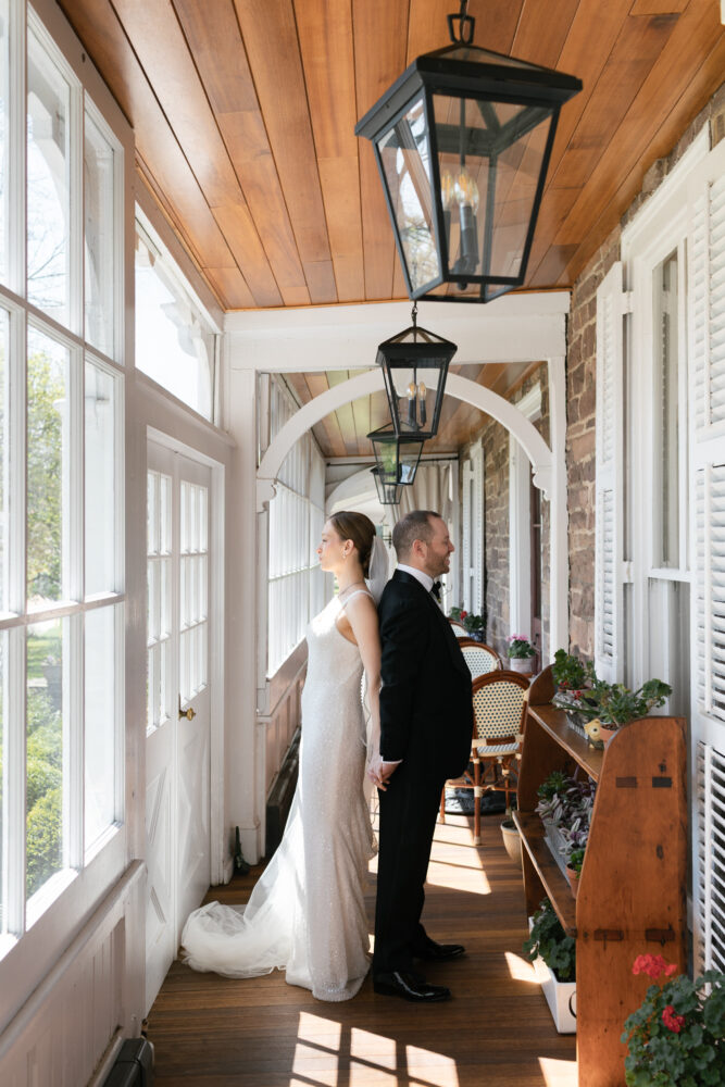 Bride in a sparkling white wedding gown and groom in a black suit stand back to back holding hands on a sunlit porch with wooden ceiling and hanging lanterns in view