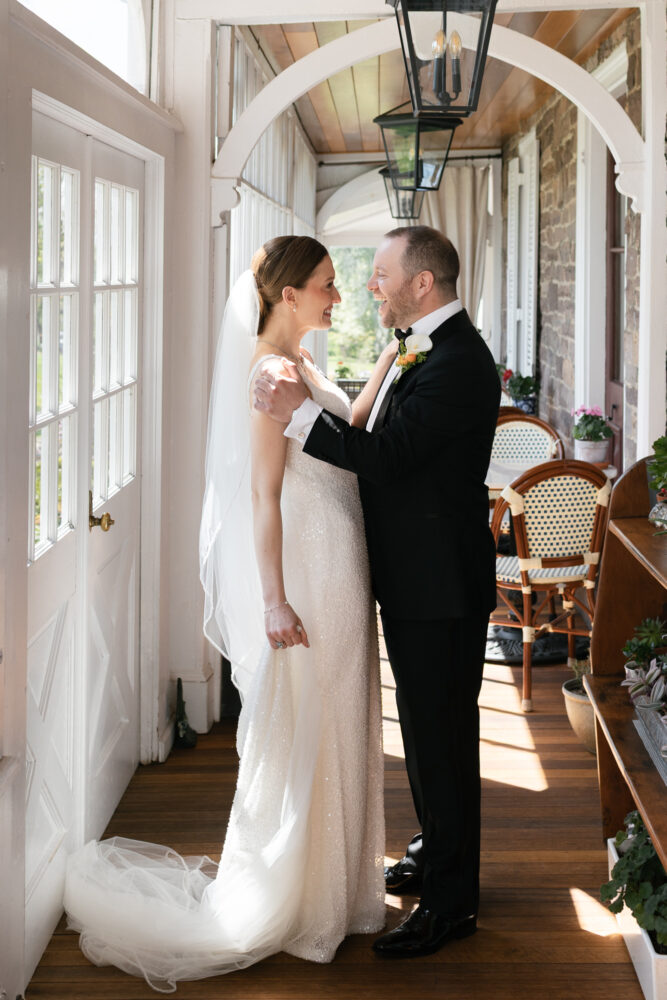 Bride in a sparkling ivory gown and veil embraces a man in a black tuxedo on a sunlit covered porch smiling at each other