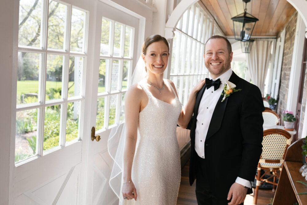 Bride in a sparkly white gown smiles beside a groom in a black tuxedo on a sunlit veranda