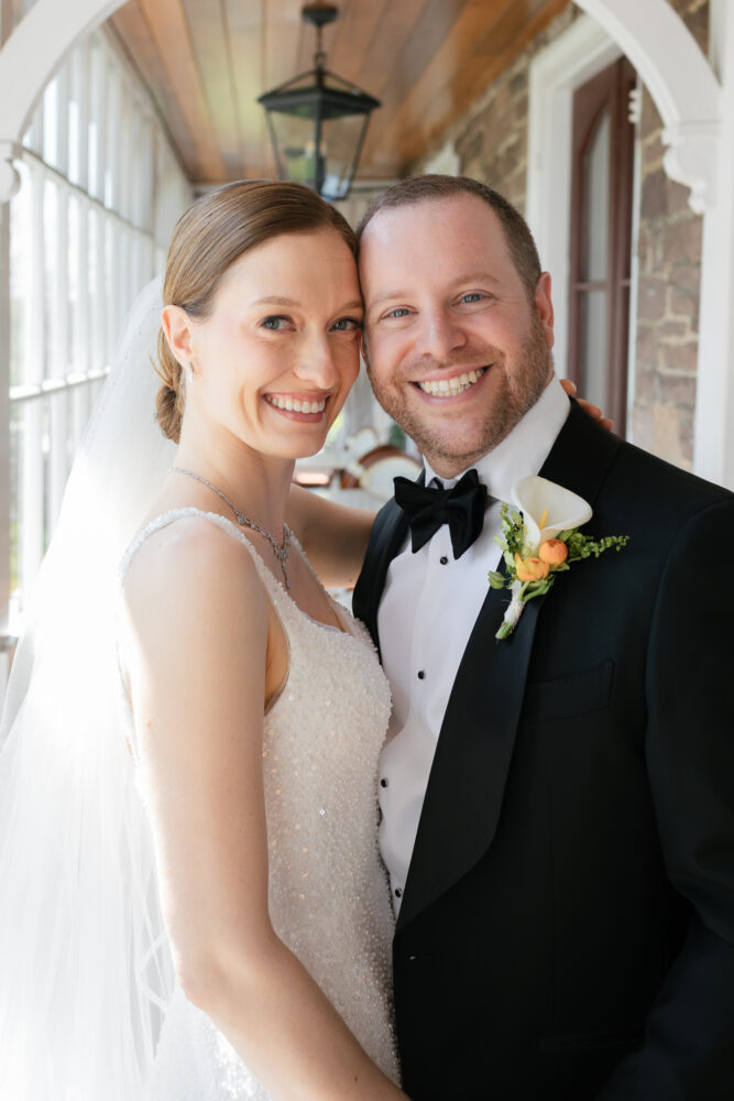 Bride in a pearl embellished wedding dress and groom in a black tuxedo smile for a close up portrait with a veil visible behind them