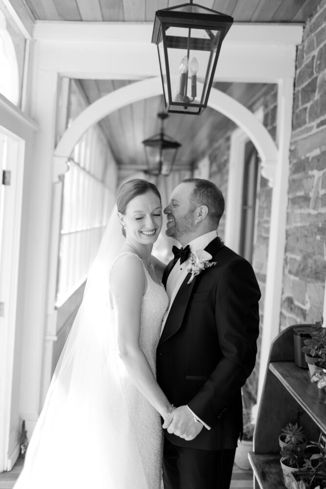 Bride and groom hold hands and smile in a arched porch hanging lanterns above them black and white wedding photo
