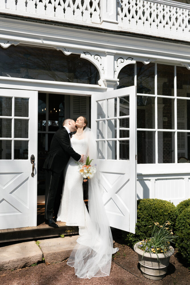 Bride in a white wedding gown and veil kissing groom in a black suit at the open white doors of a decorative building