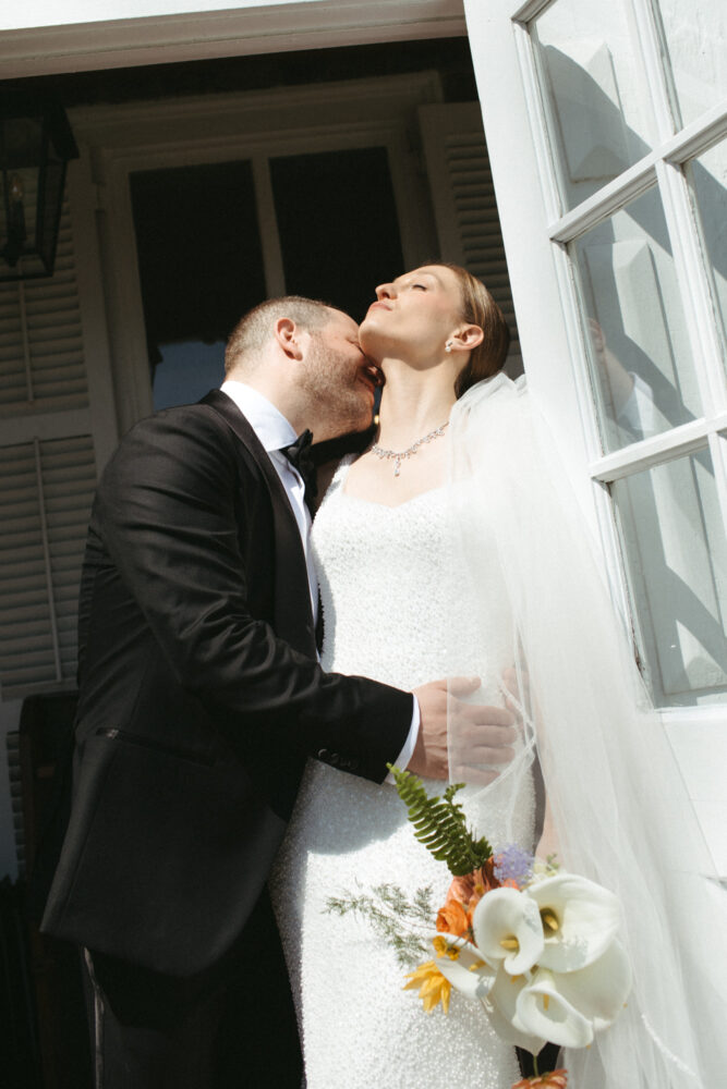 Bride in a sparkling white wedding gown and veil tilts her head back as a groom in a black tuxedo kisses her doorway in the background bouquet in hand