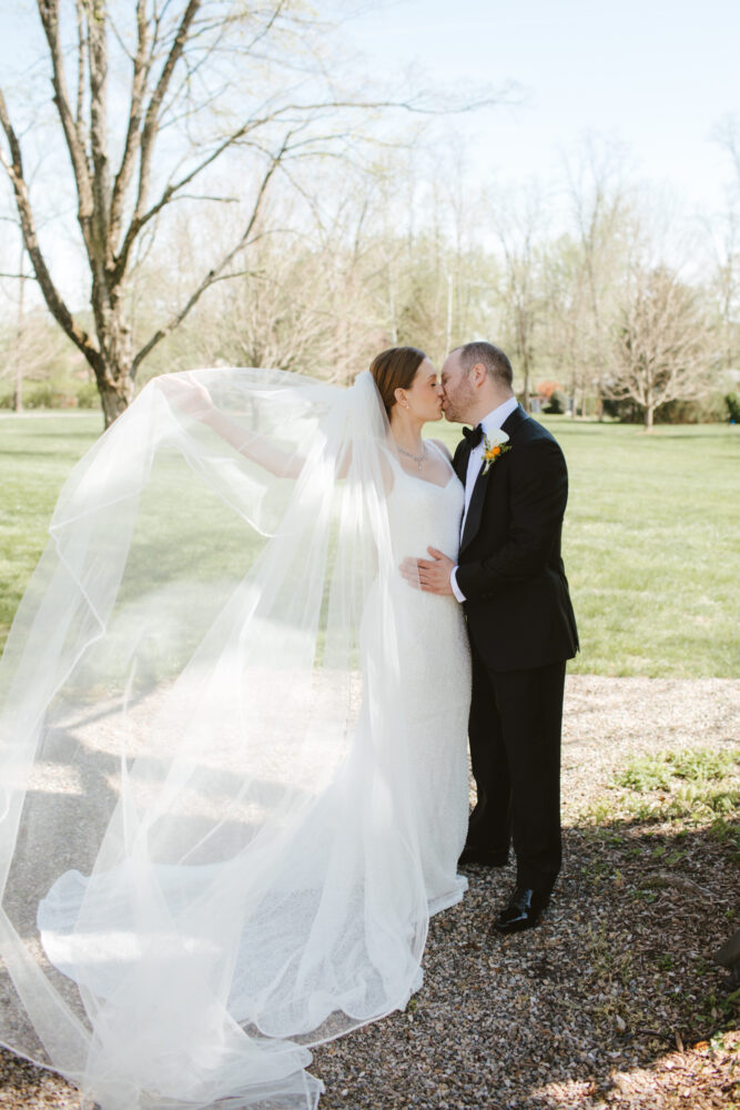 Bride and groom share a kiss outdoors the brides long veil billowing around them in a sunlit garden