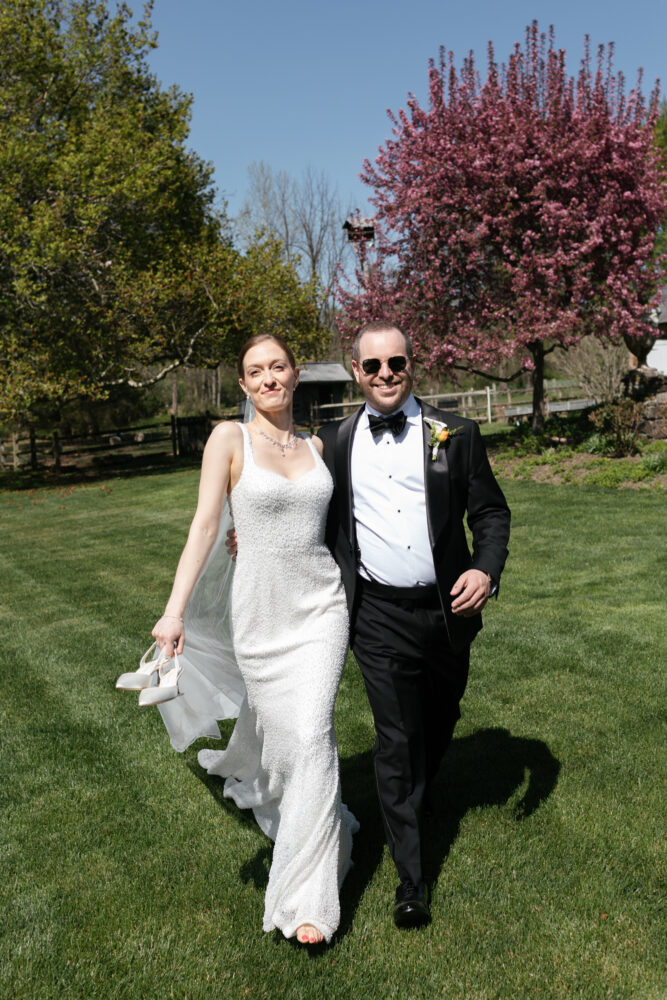Bride in a sparkling white wedding dress holding her heels walks arm in arm with a man in a black tuxedo on a sunny lawn pink flowering tree in the background