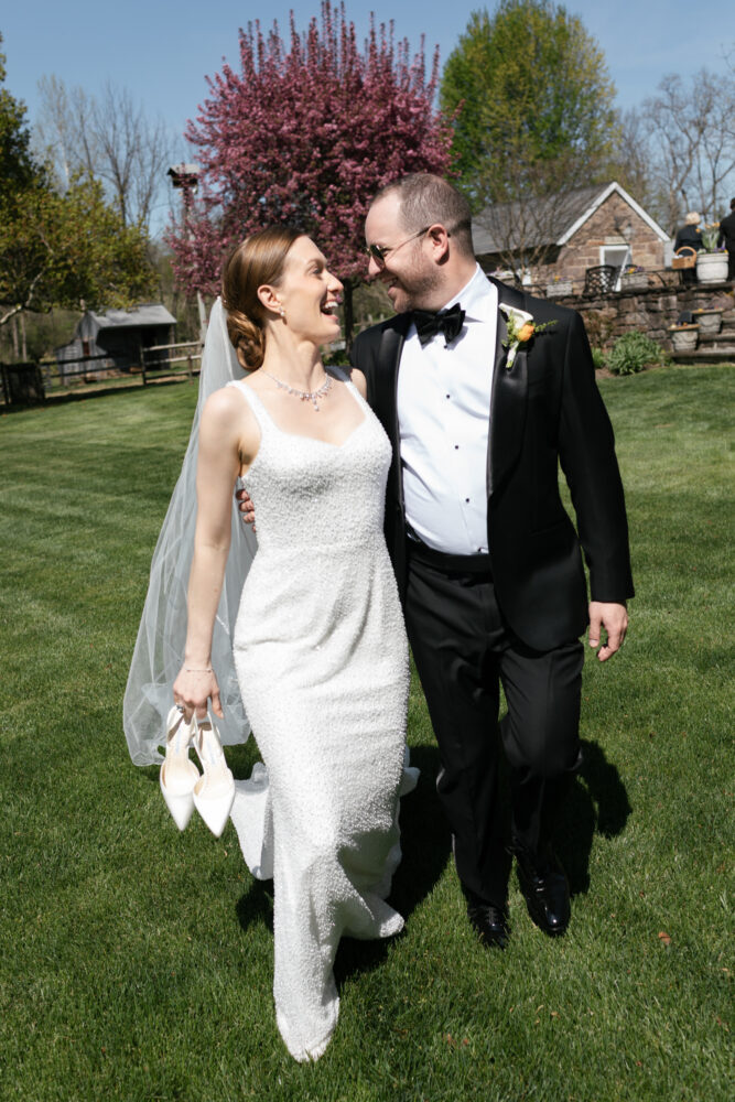 Bride and groom in wedding attire walk arm in arm across a sunny lawn the bride holding white heels in her hand