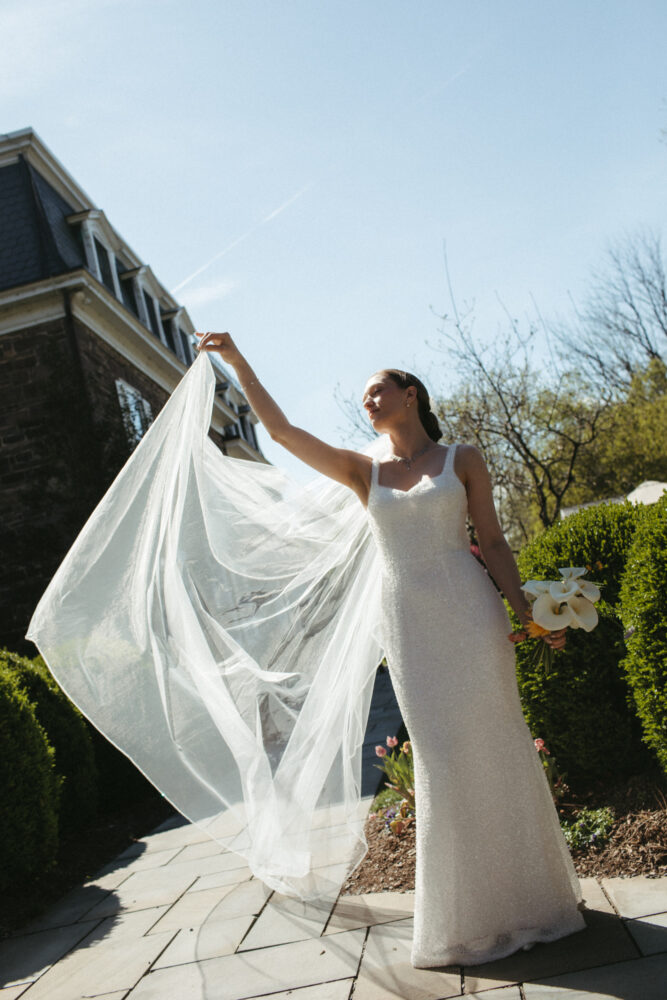 Bride in a white beaded wedding gown lifts her veil outdoors holding a bouquet of white calla lilies on her left
